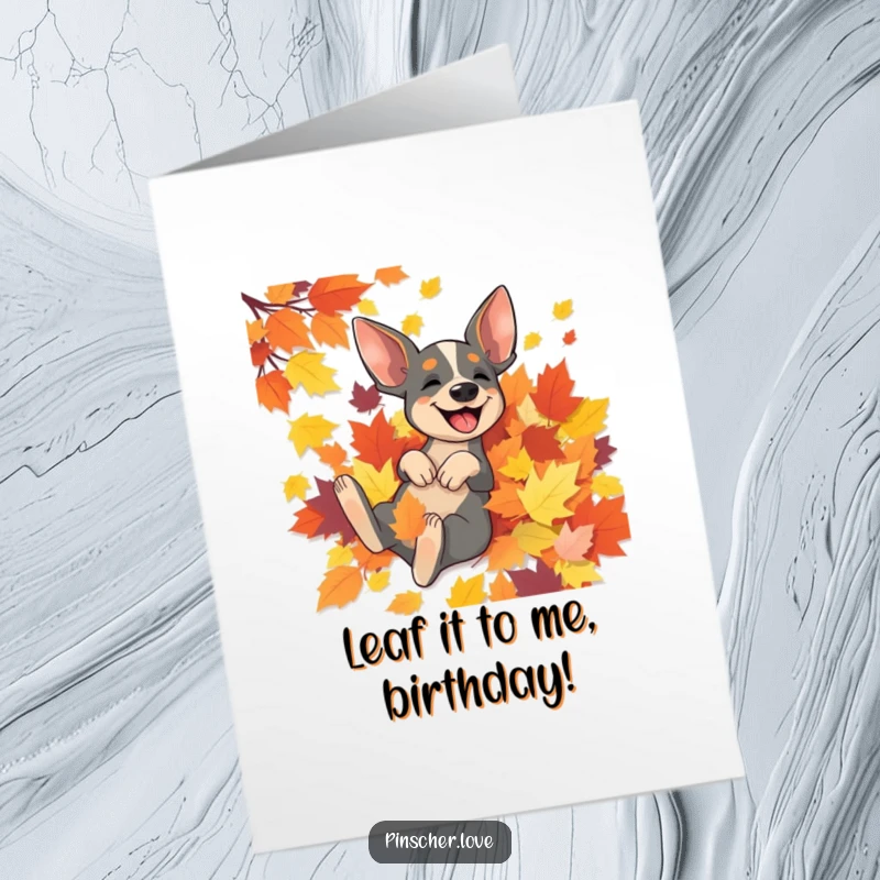 Funny free printable birthday card with a happy pinscher dog rolling gleefully in a vibrant pile of autumn leaves.